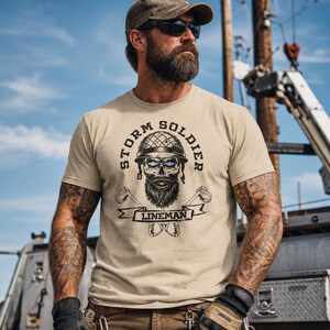 Op de afbeelding: Een beige t-shirt met korte mouwen en een grafische print van een schedel met helm en baard, met gloeiende blauwe ogen. De tekst STORM SOLDIER staat boven de schedel en LINEMAN op een banner eronder, geflankeerd door gekruiste klimijzers.