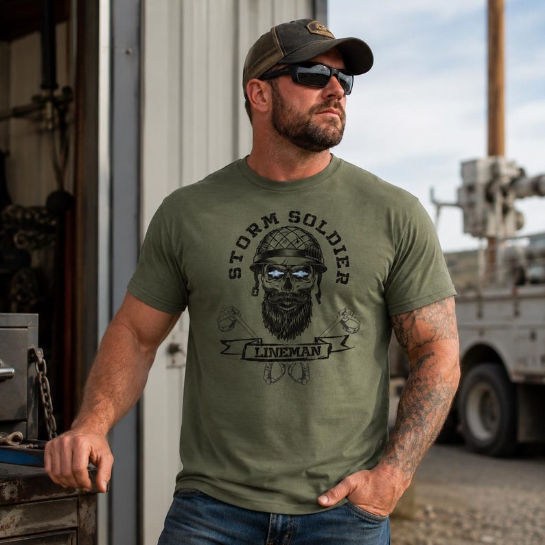 Op de afbeelding: Een olijfgroen T-shirt met korte mouwen en een zwarte grafische print van een schedel met baard en een militaire helm. De tekst STORM SOLDIER staat boven de schedel en LINEMAN op een banner eronder, met gekruiste gereedschappen.