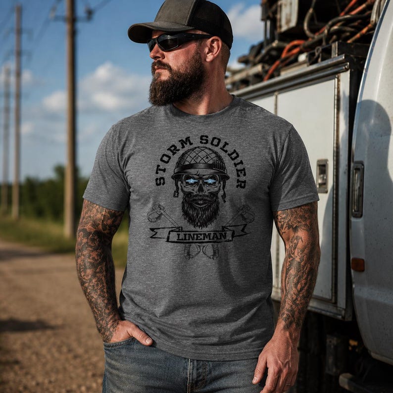 Op de afbeelding: Een antracietgrijs T-shirt met korte mouwen en een grafische print van een schedel met een helm en baard. De tekst luidt Storm Soldier Lineman, vergezeld van gekruist gereedschap. Het ontwerp is gecentreerd op de borst in zwarte inkt.
