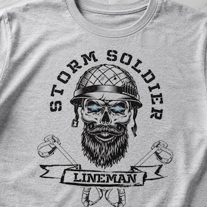 Op de afbeelding: Een lichtgrijs t-shirt met een zwarte afbeelding van een schedel met een militaire helm, een baard en een snor. De tekst STORM SOLDIER staat in een boog boven de schedel en het woord LINEMAN staat op een banner met klimuitrusting.