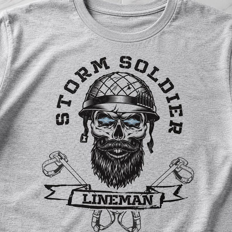 Op de afbeelding: Een lichtgrijs t-shirt met een zwarte afbeelding van een schedel met een militaire helm, een baard en een snor. De tekst STORM SOLDIER staat in een boog boven de schedel en het woord LINEMAN staat op een banner met klimuitrusting.