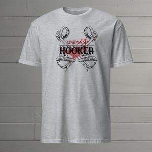 Pode incluir: Camiseta cinza com um gráfico de dois ganchos de lineman cruzados com respingos de tinta vermelha e o texto "Lineman Hooker".