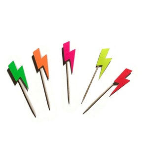 May include: Five neon coloured lightning bolt shaped paper cupcake toppers. The toppers are on wooden skewers. The colours are green, orange, pink, yellow, and red.