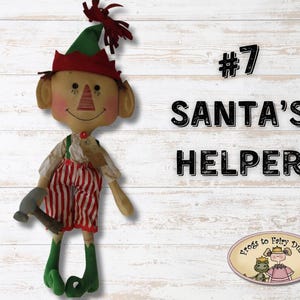 以下が含まれることがあります： 赤と白のストライプの服を着た手作りエルフ人形。緑のエルフの靴と赤いバンドのついた緑の帽子をかぶっています。人形はハンマーを持っていて、「#7 SANTA'S HELPER」とラベル付けされています。