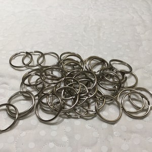 May include: A collection of silver-colored key rings, each with a circular design. The key rings are scattered on a white, patterned surface. The image showcases a bulk quantity of these metal rings.