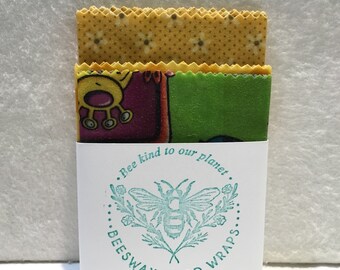 May include: A set of three beeswax food wraps with a yellow floral pattern, a colorful pattern with a cartoon character, and a white label with a bee and the text "Bee kind to our planet. Beeswax Food Wraps."