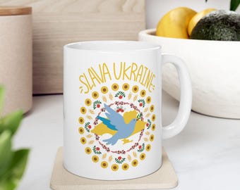 Op de afbeelding: Witte keramische mok met de tekst "SLAVA UKRAINE" in gele letters. Het ontwerp bevat een blauwe duif, een geel-blauwe kaart, zonnebloemen en bloemmotieven. De mok staat op een beige onderzetter.