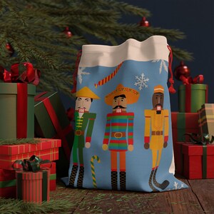 May include: A blue drawstring gift bag with three nutcracker figures in colourful outfits. The bag has a white top with snowflakes and red drawstrings. The background includes wrapped gifts and a Christmas tree.