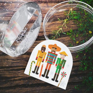 Puede incluir: Una tarjeta navideña festiva con tres figuras de cascanueces ilustradas con trajes coloridos, incluyendo un sombrero y un gorro cónico. La tarjeta está sobre una superficie de madera, con un recipiente de vidrio transparente y vegetación en el fondo.