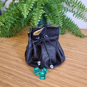 May include: A black, textured leather dice bag with a drawstring closure and white bead accents. The bag is open, revealing a gold interior. Three green dice with white numbers are in front of the bag. A fern plant is in the background.