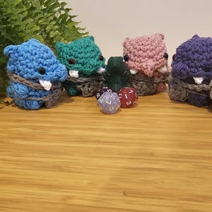 Puede incluir: Una colección de figuras de monstruos de ganchillo hechas a mano en azul, verde, rosa y morado. Cada monstruo tiene colmillos blancos, ojos negros y está envuelto con hilo gris. Varios dados están frente a los monstruos.