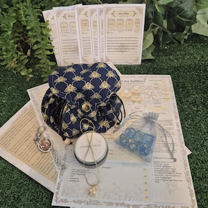 May include: A collection of tabletop gaming accessories, including a navy blue drawstring bag with a gold bee pattern, dice, a necklace, and printed game cards. The items are arranged on a green surface.