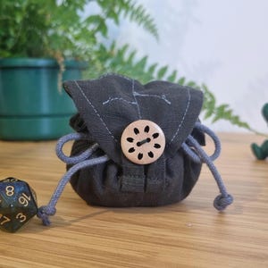 以下が含まれることがあります： 木製のボタンが付いた小さな黒い巾着袋。袋は柔らかく、質感のある生地で作られており、灰色の紐が付いています。袋は木製の表面の上に置かれています。