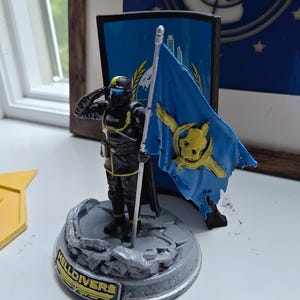 May include: A Helldivers figurine featuring a soldier in black and yellow armor saluting while holding a blue flag with a yellow skull emblem. The figure stands on a gray, rocky base with the text HELLDIVERS printed on the front edge in yellow.