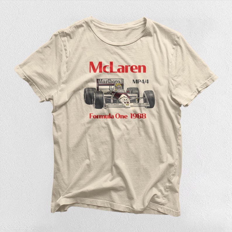 May include: A beige t-shirt featuring a red McLaren logo, a red "MP4/4" text, a red "Marlboro" text, and a black and white image of a Formula One race car. The text "Formula One 1988" is printed below the image.