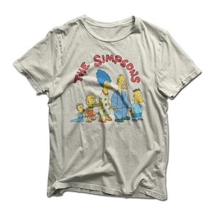 Könnte beinhalten: Cremefarbenes T-Shirt mit einer Grafik der Familie Simpson. Das Design zeigt Homer, Marge, Bart, Lisa und Maggie, mit dem Text "The Simpsons" in Rot. Das Shirt besteht aus einem weichen, bequemen Material.