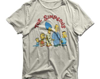 May include: Cream-coloured t-shirt with a graphic of the Simpsons family. The design features Homer, Marge, Bart, Lisa, and Maggie, with the text "The Simpsons" in red. The shirt is made of a soft, comfortable material.
