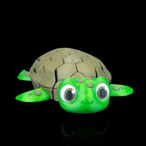 May include: A 3D-printed green turtle figurine with a light brown segmented shell. The turtle has large, expressive eyes and small brown spots on its head and flippers. The figurine is set against a black background.