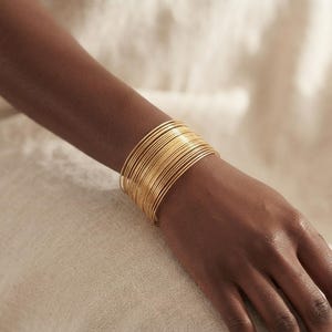 Peut inclure: Un large bracelet manchette composé de nombreuses fines bandes circulaires en fil doré empilées pour créer une pièce métallique unique, continue et texturée qui entoure le poignet avec une finition dorée uniforme, polie et réfléchissante.