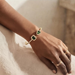 Könnte beinhalten: Ein offener Goldarmreif mit einem quadratisch geschliffenen grünen Edelstein und einem birnenförmigen grünen Edelstein, beide umgeben von kleinen weißen Steinen. Das Armband ist ebenfalls mit kleinen weißen Steinen besetzt. Zwei schlichte Goldringe.