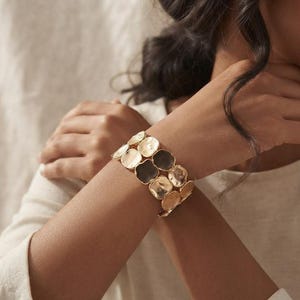 Può includere: Un bracciale a fascia larga composto da molteplici dischi metallici circolari interconnessi. I dischi presentano una superficie martellata e testurizzata con una finitura dorata riflettente, creando un aspetto stratificato sul polso.