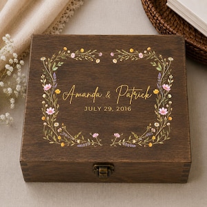 May include: A square dark brown wooden box with a metal latch, featuring a printed floral wreath design in pink, yellow, and green. The center text reads Amanda & Patrick and the date JULY 29, 2016 in a gold-colored script and block font.