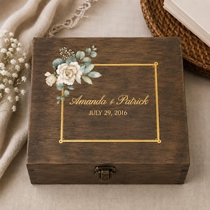 May include: A square dark brown wooden keepsake box with a metal latch. The lid features a gold rectangular frame, a floral illustration of white roses and eucalyptus leaves, and the printed text Amanda & Patrick JULY 29, 2016.