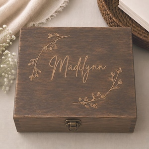 May include: A square dark brown wooden keepsake box featuring a hinged lid with a metal latch. The top surface is engraved with the name Madilynn in a cursive script, framed by two delicate, mirrored floral branch designs on either side.