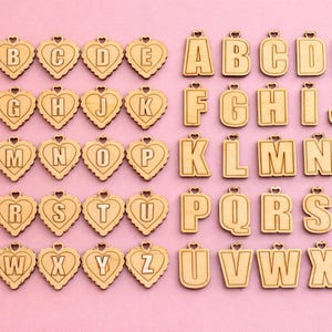 May include: Wooden alphabet letters and heart-shaped pendants with letters, arranged on a pink background. The letters are in a sans-serif font. The heart pendants have a scalloped edge and a small heart at the top for hanging.