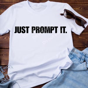 Op de afbeelding: Een wit T-shirt met korte mouwen en de zwarte tekst JUST PROMPT IT. gecentreerd op de borst. Het shirt wordt getoond naast een zonnebril met een bruin montuur en lichtblauwe spijkerstof, wat zorgt voor een eenvoudig en casual ontwerp.