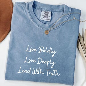 May include: A blue Comfort Colors t-shirt with the words "Live Boldly, Love Deeply, Lead With Truth" in white script. A gold necklace with a pendant rests on the shirt. The shirt is folded.