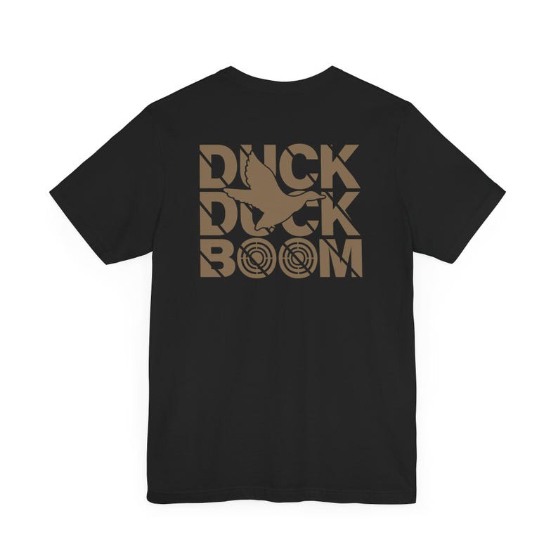 Puede incluir: Camiseta negra con un estampado gr&aacute;fico marr&oacute;n de un pato volando a trav&eacute;s de un objetivo con el texto "DUCK BOOM" impreso en una fuente de bloque.
