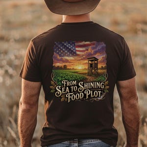Puede incluir: Camiseta marrón con un gráfico que presenta una bandera estadounidense, un puesto de caza y el texto "From Sea to Shining Food Plot". El diseño incluye una puesta de sol sobre un campo.