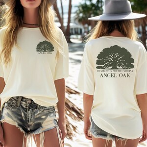May include: Cream-colored t-shirt with a green Angel Oak tree graphic on the front and back. The back of the shirt also features the text "Charleston South Carolina". The shirt is a casual, short-sleeved design.
