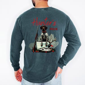 May include: Dark teal long-sleeve shirt with a graphic on the back. The graphic features a hunting dog, a rifle, a duck, and the words "Hunter's Pride" in red script. The shirt is made of a soft, comfortable material.