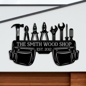May include: Black metal sign featuring tools like a hammer, pliers, and wrench above a tool belt. The sign reads "THE SMITH WOOD SHOP EST. 2012". The sign is mounted on a white wall above a wooden garage door.