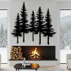 Op de afbeelding: Zwarte metalen silhouet wandkunst die een groep van vijf dennenbomen met een open haard en houtblokken op de voorgrond afbeeldt. De open haard heeft een brandend vuur en de kamer is ingericht met witte banken en een salontafel.