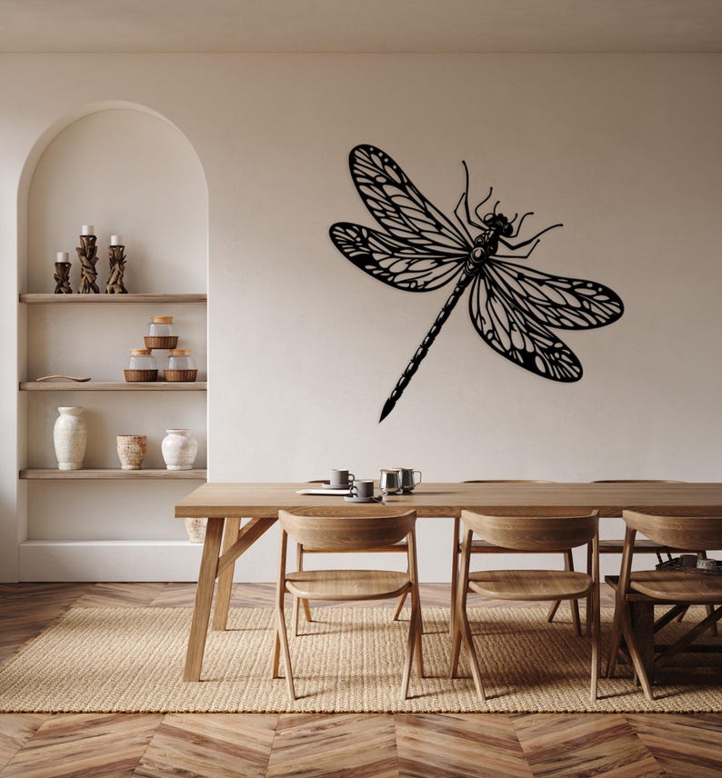 May include: A black metal dragonfly wall decor with intricate details hangs above a wooden dining table with chairs. The table is set with cups and saucers.