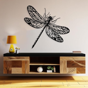 May include: Black metal dragonfly wall decor with intricate details, mounted on a white wall above a wooden shelf with a lamp and plants.