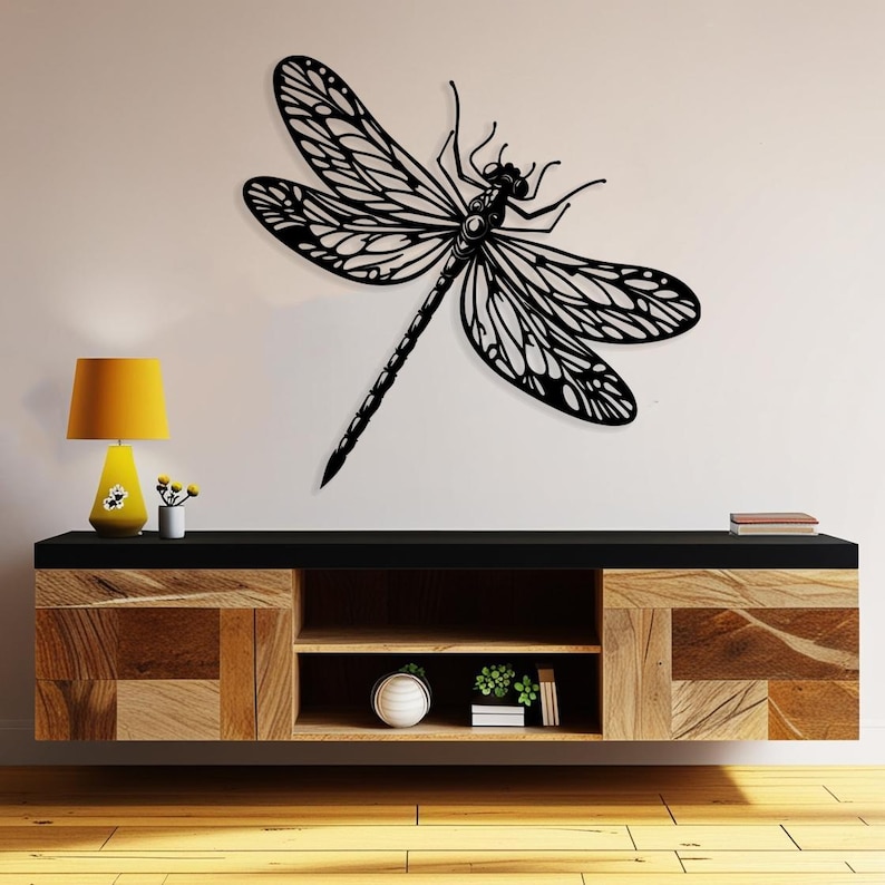 May include: Black metal dragonfly wall decor with intricate details, mounted on a white wall above a wooden shelf with a lamp and plants.