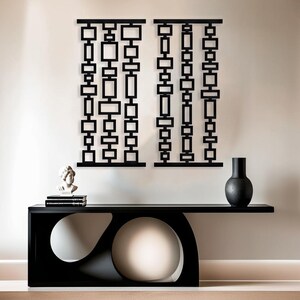 May include: Two black metal wall sculptures with geometric shapes, hanging on a white wall above a black console table with a white round object in the center.