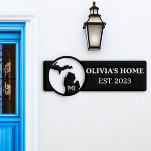 以下が含まれることがあります： 「OLIVIA'S HOME EST. 2023」と書かれた黒い金属製の家の看板。ミシガン州のシルエットと「MI」の文字が入った円形のデザインです。看板の上には黒い屋外用ランタンが取り付けられています。