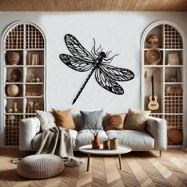 May include: A black and white silhouette of a dragonfly with intricate wing details is displayed on a white wall. The dragonfly is centered in the image and is the main focal point. The wall is in a living room setting with a couch, coffee table, and decorative items.