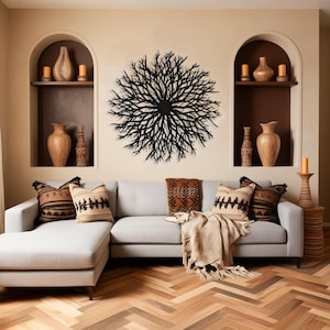 May include: A light gray sectional sofa with a throw blanket and pillows in a living room with a black metal wall sculpture of a tree branch design. The room has a light beige wall with two arched niches with brown pottery vases and candles.