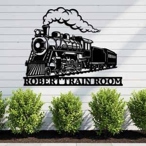 May include: Black metal train wall art with the text "ROBERT TRAIN ROOM". The train is a detailed silhouette of a classic steam engine with a billowing cloud of smoke. The artwork is mounted on a white wooden wall with green bushes in the foreground.