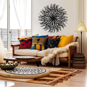 May include: A white couch with colorful patterned throw pillows and a fluffy white blanket. A black metal wall sculpture resembling a tree branch is hanging on the wall behind the couch. A wooden coffee table with a plate on it sits in front of the couch. A round, patterned cushion sits on the floor in front of the couch.