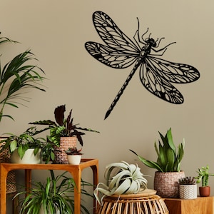 May include: Black metal dragonfly wall decor with intricate details. The dragonfly is mounted on a light beige wall with potted plants in the foreground.