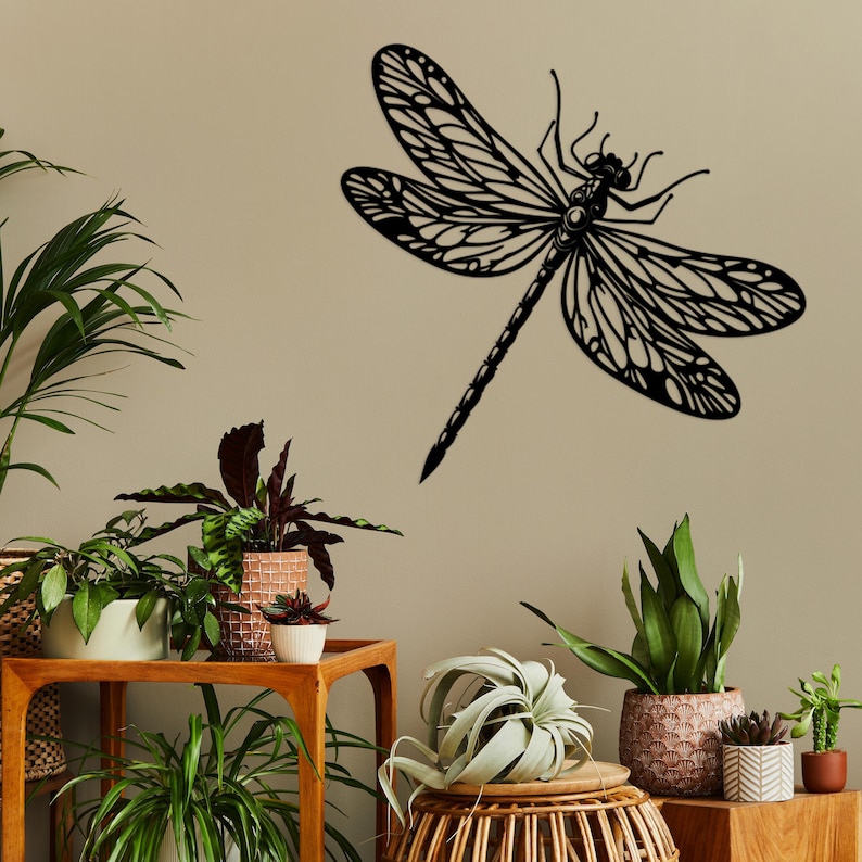 May include: Black metal dragonfly wall decor with intricate details. The dragonfly is mounted on a light beige wall with potted plants in the foreground.