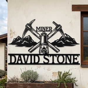 Può includere: Arte da parete in metallo nero a tema minatore. Il design include picconi incrociati, una lanterna e sagome di montagne, con la parola "MINER" sopra e "DAVID STONE" sotto.