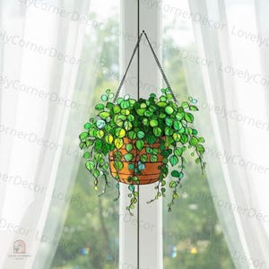 Op de afbeelding: Een gebrandschilderde hangende plantenbak met een ketting, gevuld met groene bladplanten. De plantenbak heeft een warme bruine kleur en hangt voor een raam met witte gordijnen. De tekst "LovelyCornerDecor" is zichtbaar.